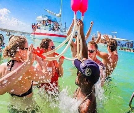 Personas en agua turquesa poco profunda levantan un embudo rosa con tubos blancos; al fondo, un catamarán