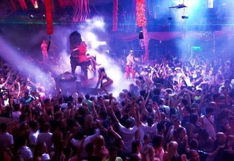 Sala llena de público frente al escenario, con luces moradas y humo