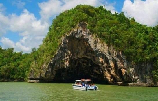 Lancha con pasajeros frente a un peñón cubierto de vegetación con una gran cueva a nivel del agua