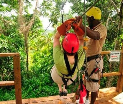 Persona con casco rojo y arnés cuelga del cable junto a un guía en una plataforma de tirolina