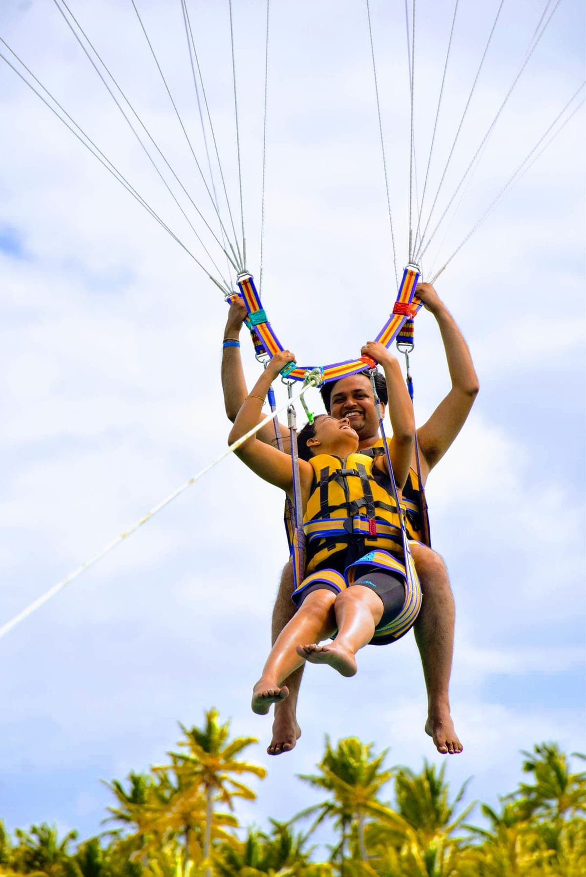 Pareja sonriente con chalecos salvavidas en el arnés de parasailing, con palmeras debajo