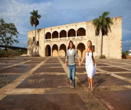 Pareja caminando de la mano por una plaza adoquinada frente al Alcázar de Colón, con palmeras