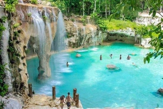 Cascadas caen por una pared de roca a una laguna turquesa con bañistas; una pareja baja por un sendero.