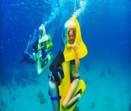 Turista saluda desde un scooter submarino amarillo con casco burbuja; detrás, otro scooter y un buzo de apoyo
